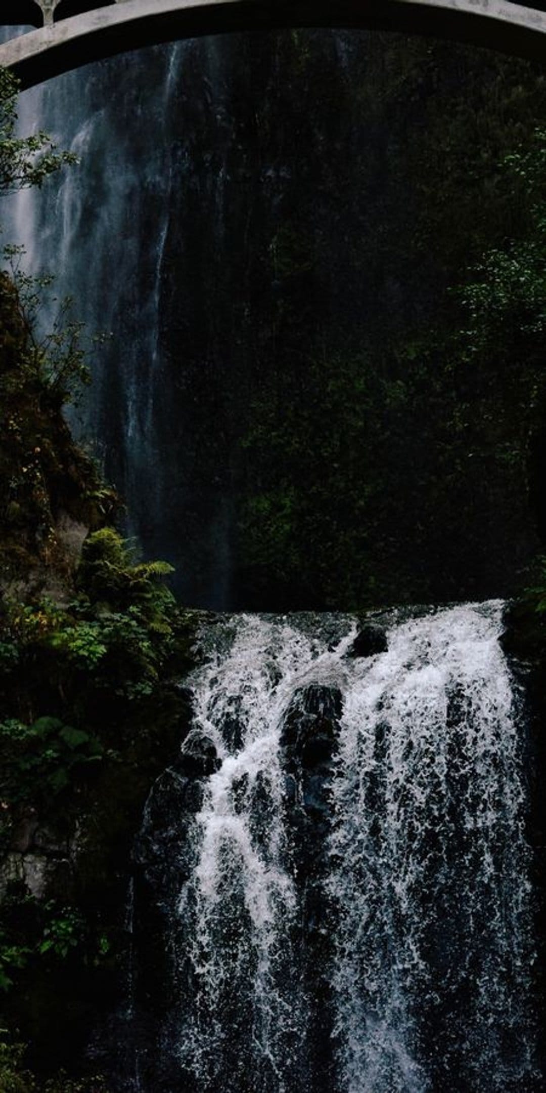 Tiered white waterfall rushing over mossy black rocks beneath a stone arch bridge in a lush forest setting.