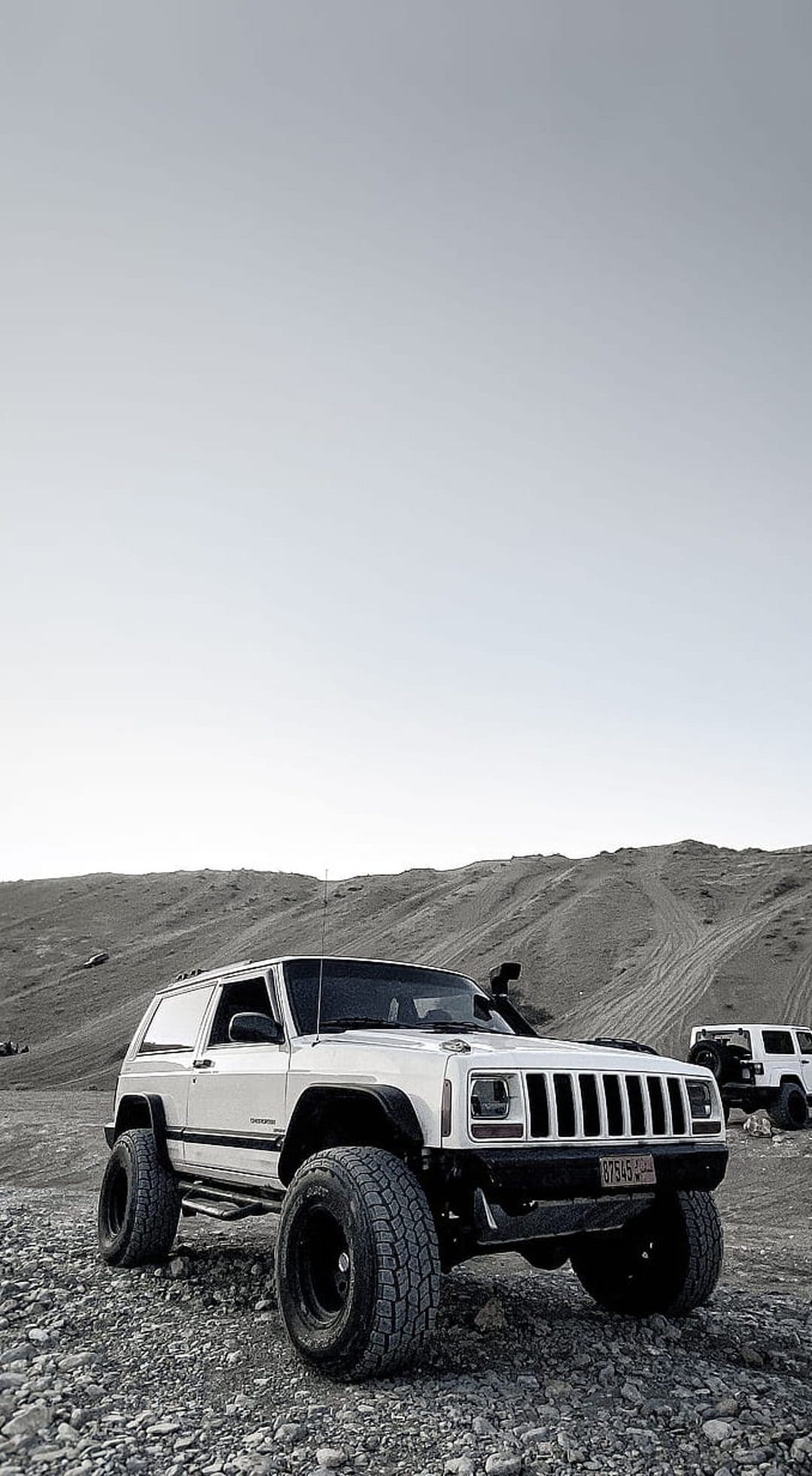 White two-door Jeep Cherokee with oversized tires parked on gravel terrain with sandy hills in the background.