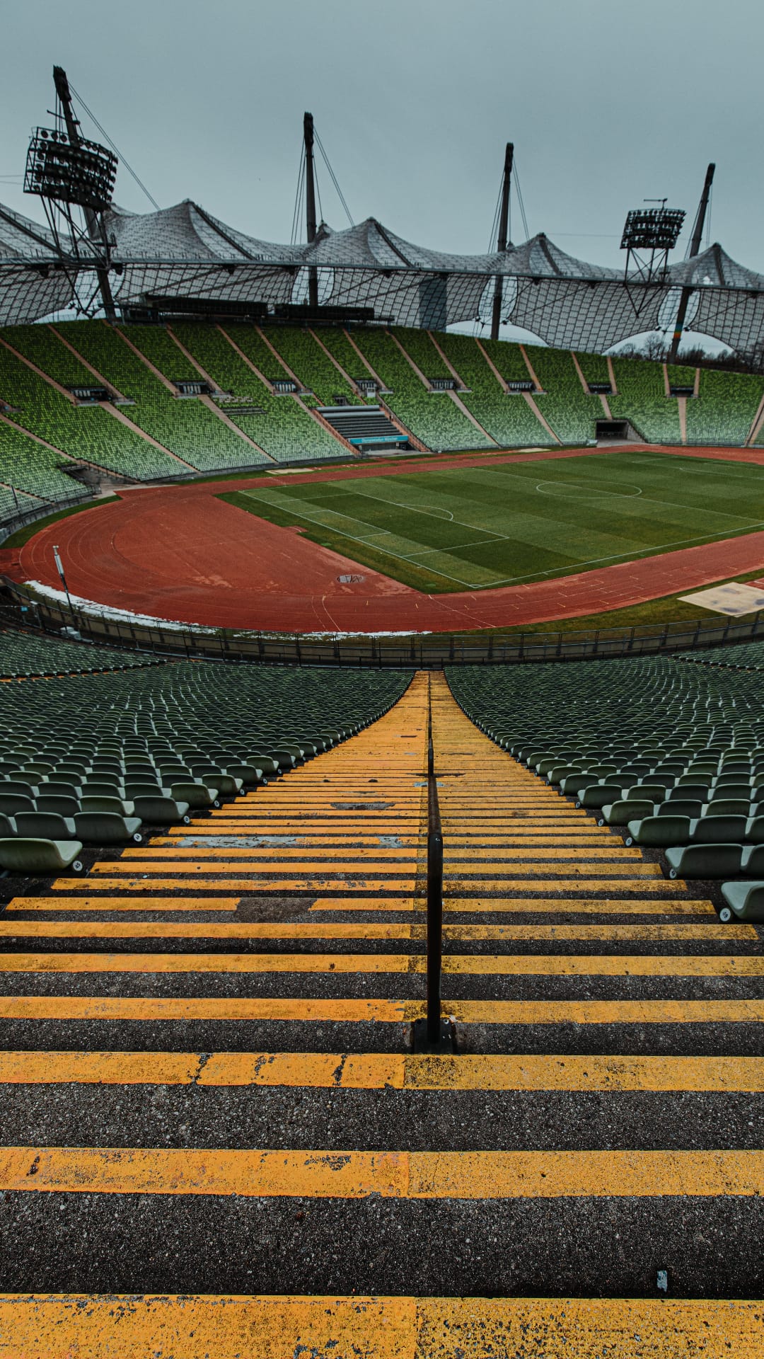 Yellow and black striped concrete steps lead toward rows of empty green seats and a red athletics track under a roof.