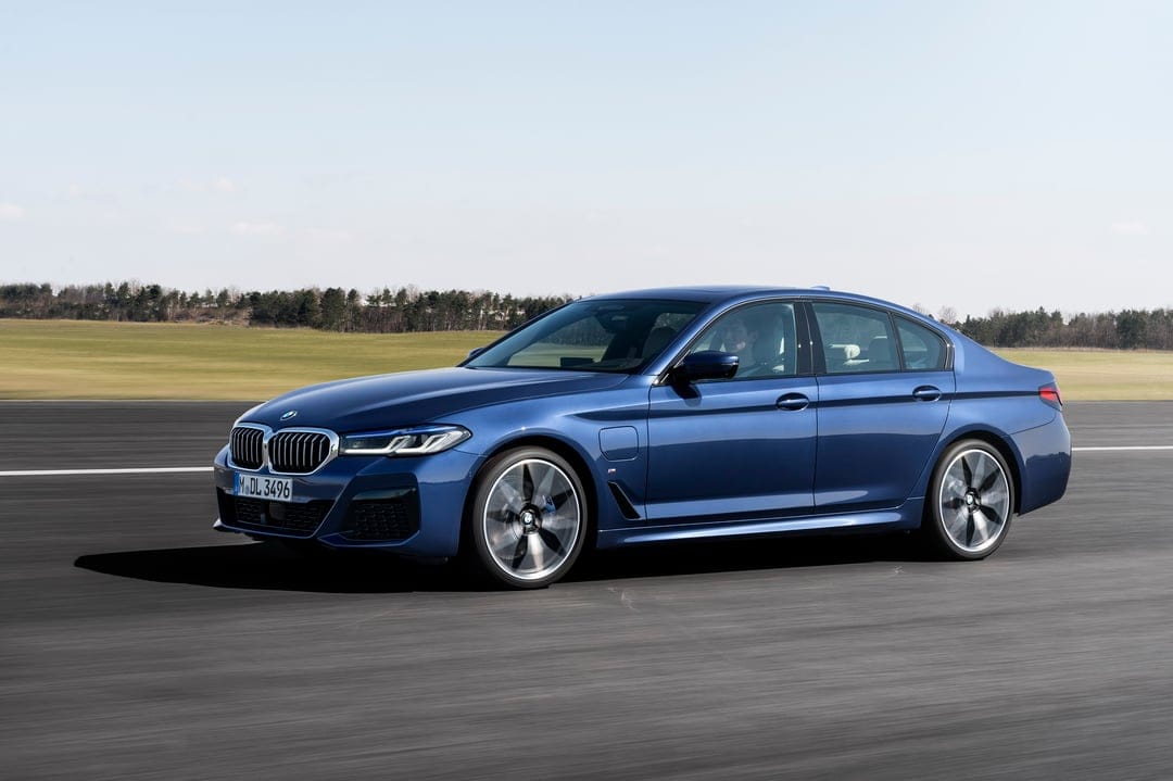 Blue BMW sedan driving on a track with motion-blurred wheels against a background of green fields and clear sky.