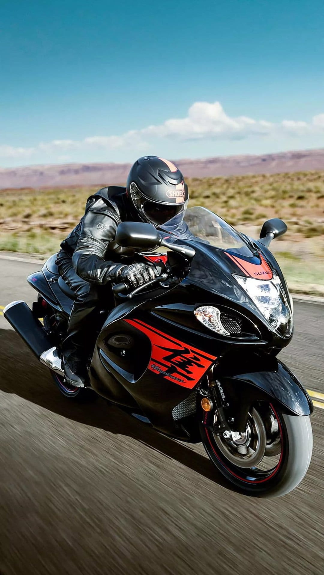 Suzuki Hayabusa rider in black leathers speeding on a desert road under a blue sky with distant mountains.