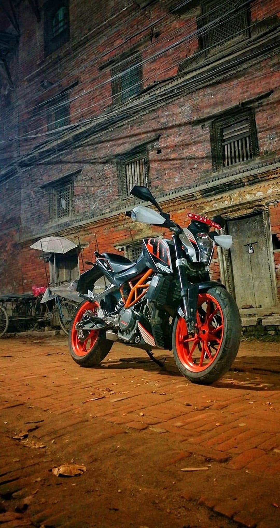KTM Duke motorcycle with orange wheels parked on a cobblestone street by a brick building at night.