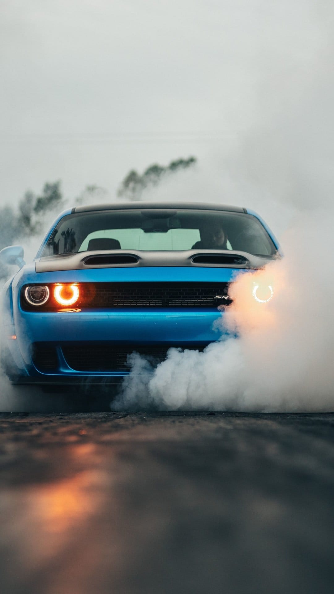 Blue muscle car front view during a burnout with heavy white smoke and glowing orange halo headlights on asphalt.