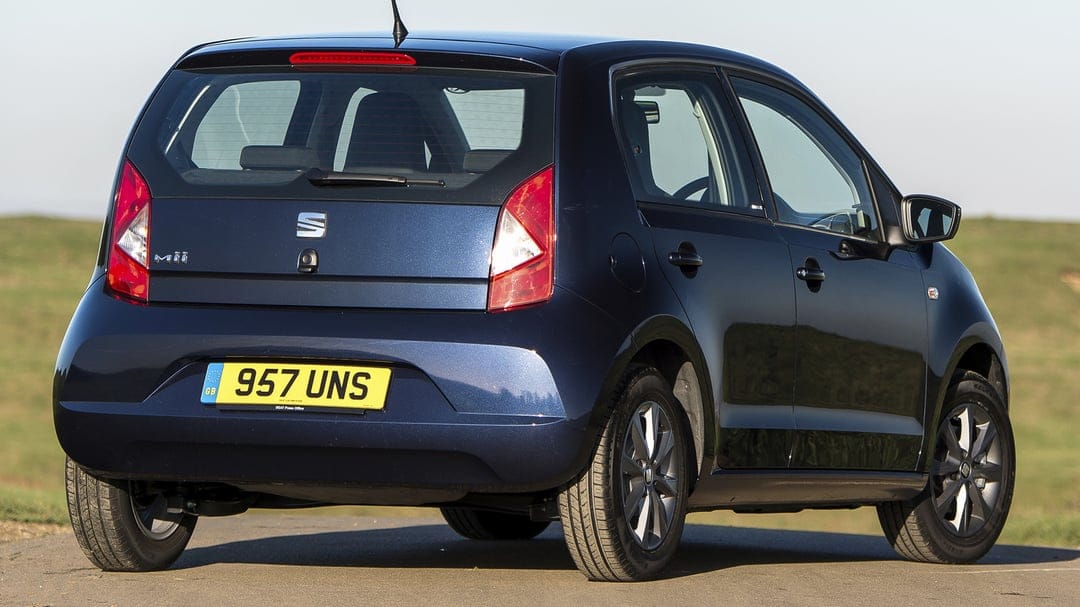 Dark blue SEAT Mii hatchback parked on a quiet road with a blurred green field and clear sky in the background.