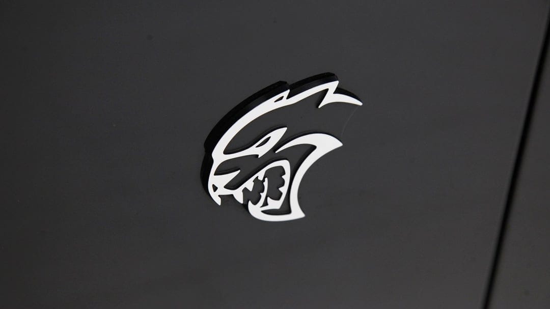 White Dodge Hellcat logo centered on a dark matte grey surface with subtle shadows and clean lines.