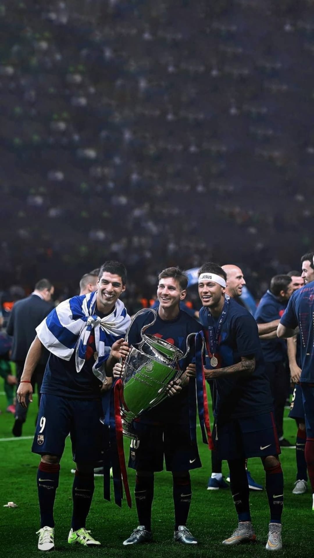 Messi, Suárez, and Neymar hold a large gold trophy while wearing striped Barcelona jerseys in a packed stadium.