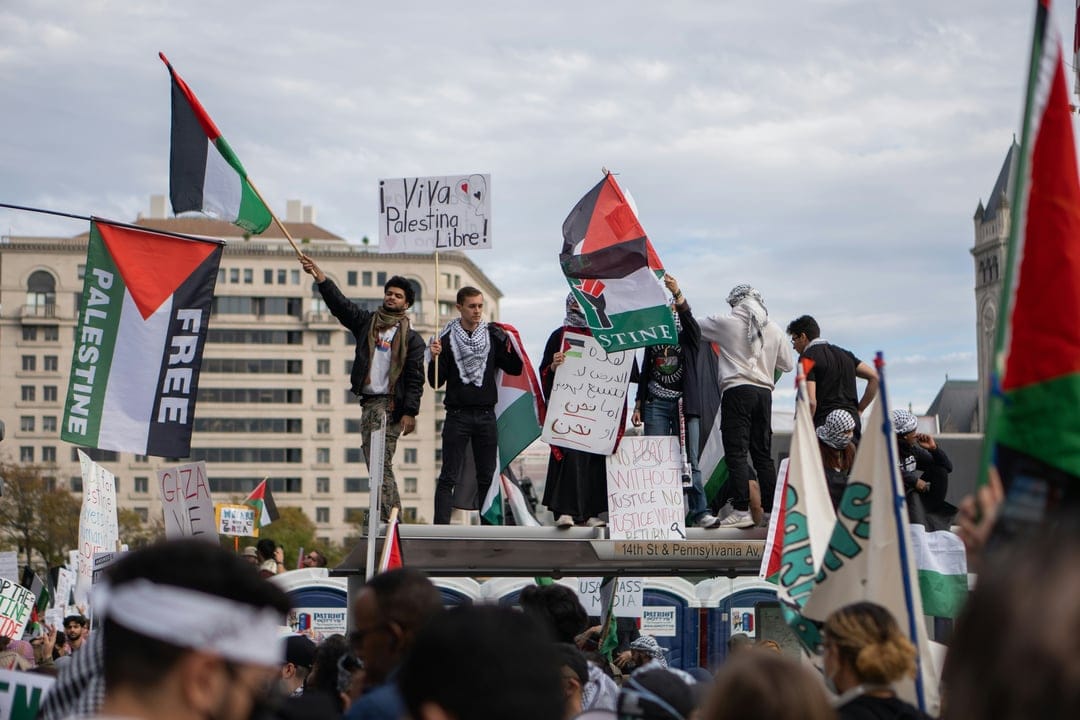 Palestinian flags and a Viva Palestina Libre sign held by a crowd of protesters near an urban building.