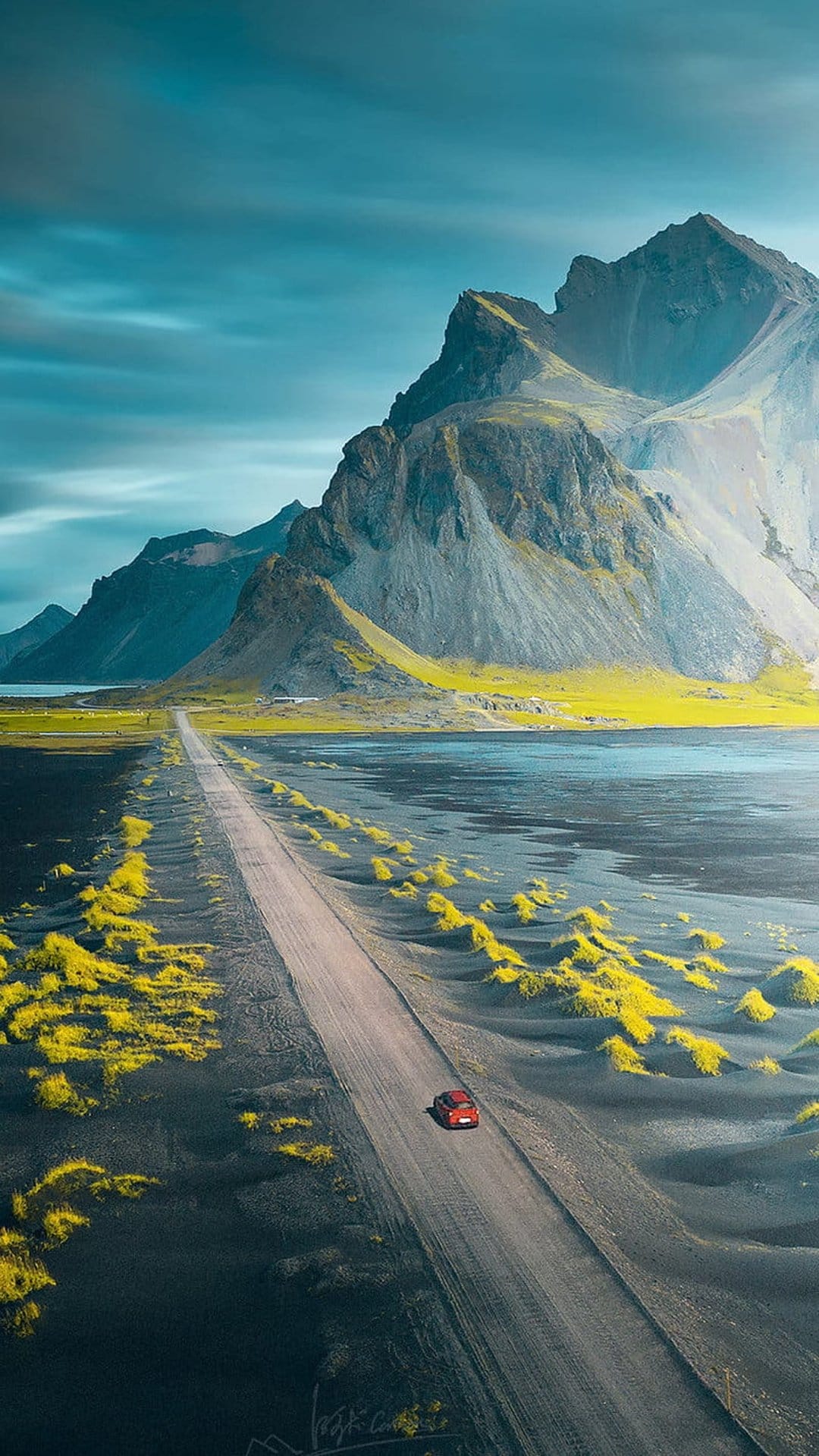 Red vehicle drives on a gravel road between green mossy fields and jagged volcanic peaks under a cloudy sky.