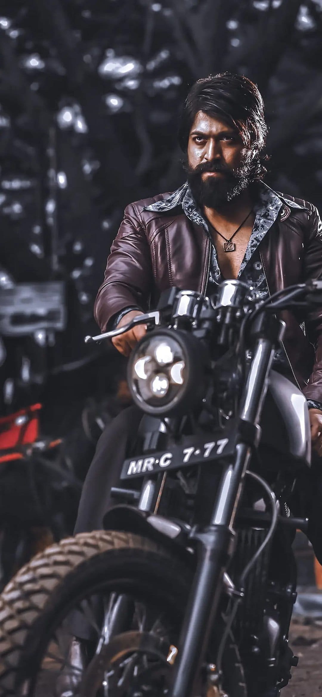 Rocky Bhai with a thick beard and leather jacket sits on a black motorcycle against a dark, cinematic backdrop.