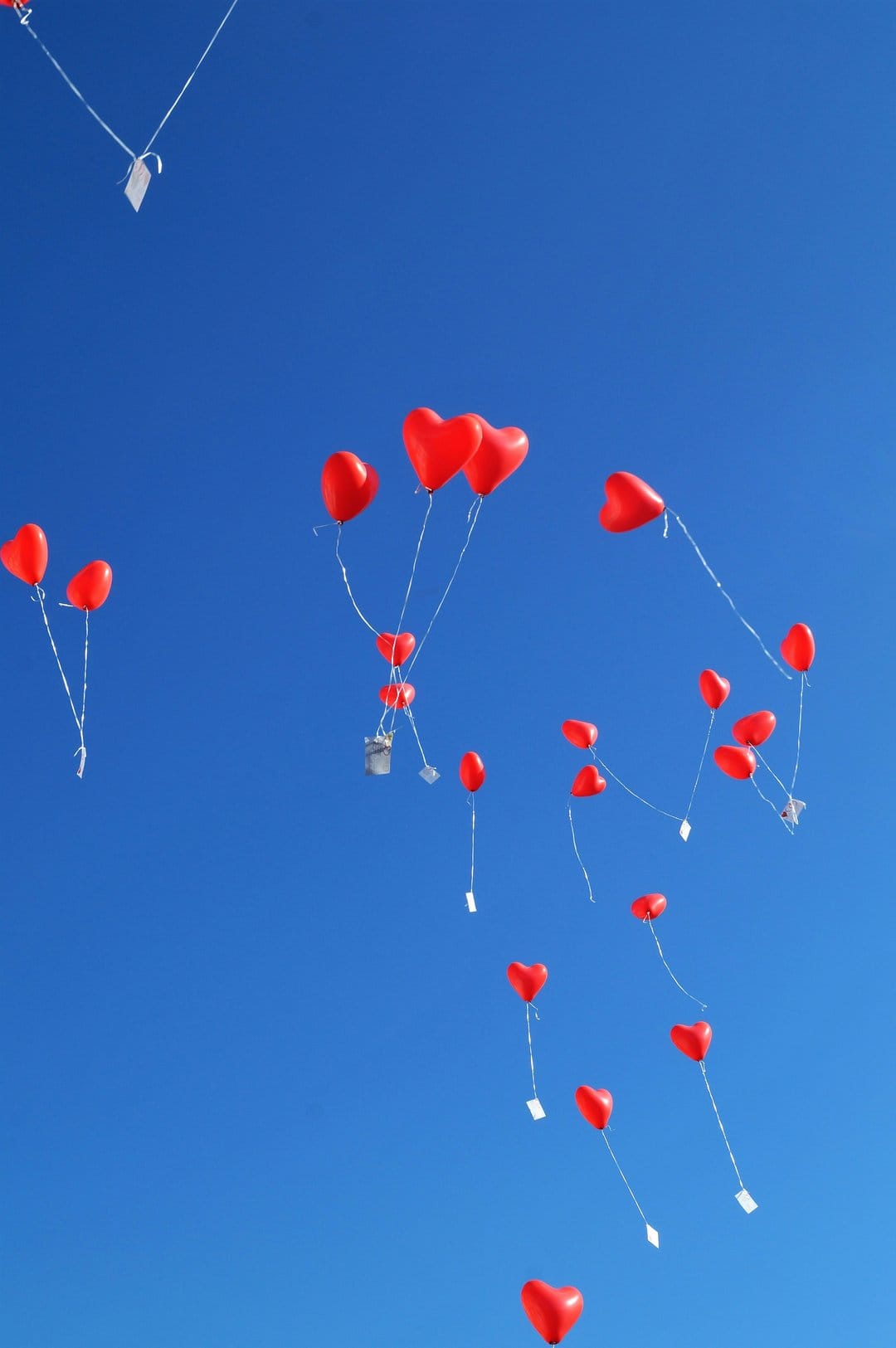 Red heart-shaped balloons with white strings ascend through a cloudless bright blue sky in bright daylight.