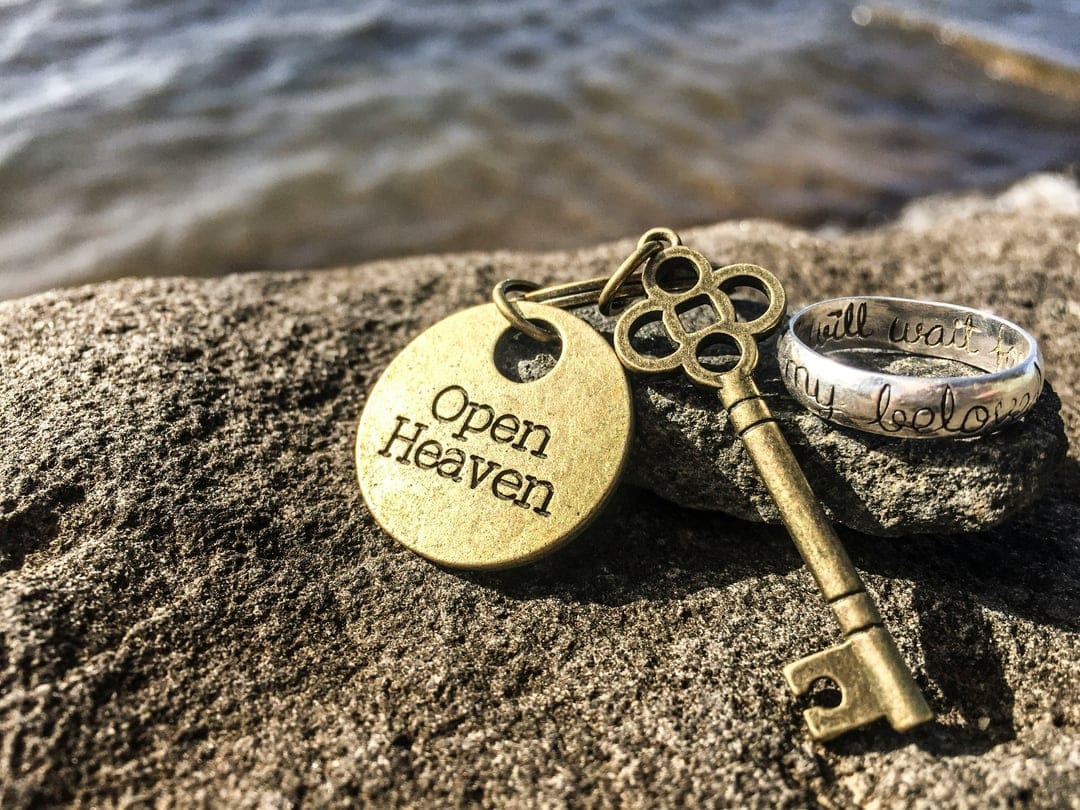 Brass key charm and silver engraved ring resting on grey textured rocks with blurred blue water in the background.