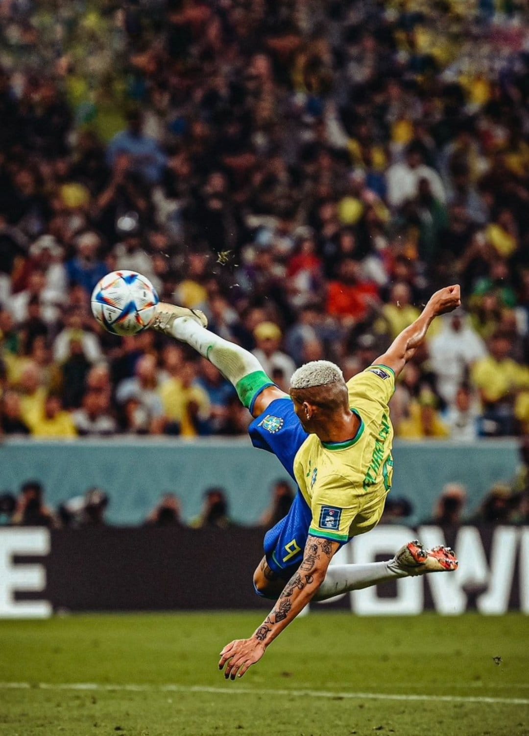 Richarlison mid-air performing a bicycle kick while wearing a yellow Brazil jersey during a soccer match.