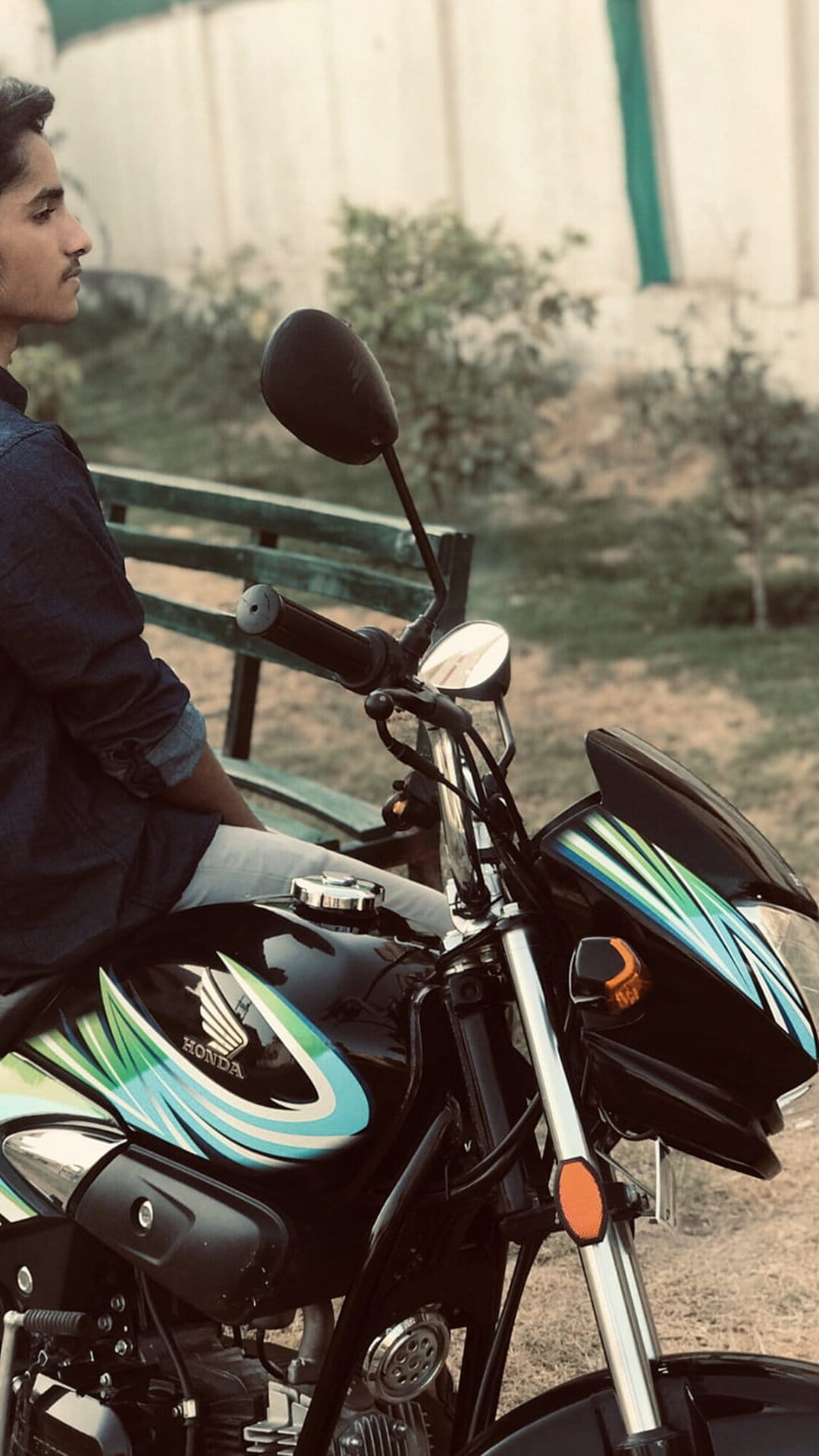 Young man in a denim shirt sits on a black Honda motorcycle with green graphics in a sunlit outdoor park.