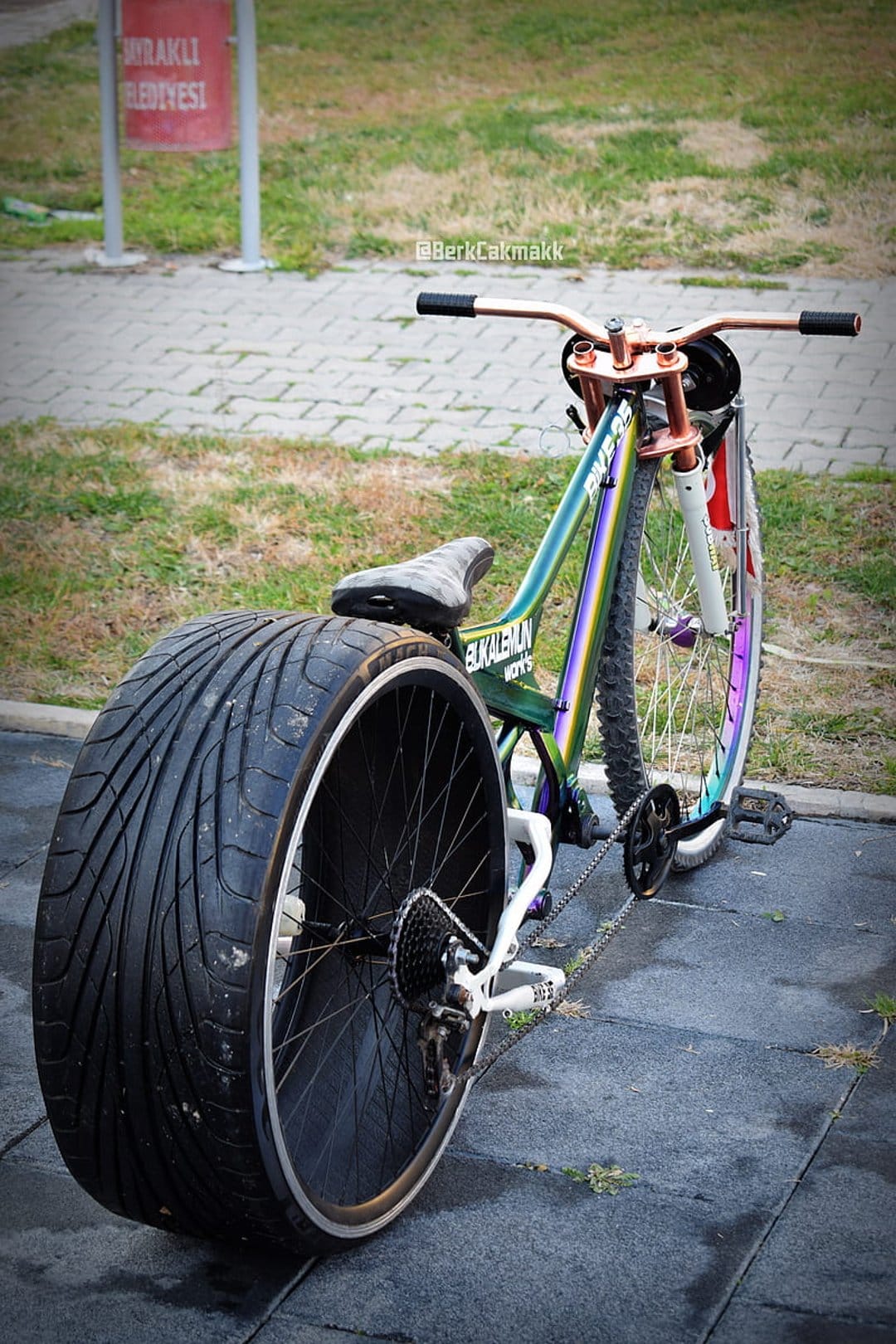 Custom iridescent bicycle with a huge car tire rear wheel and copper handlebars on a paved urban path.