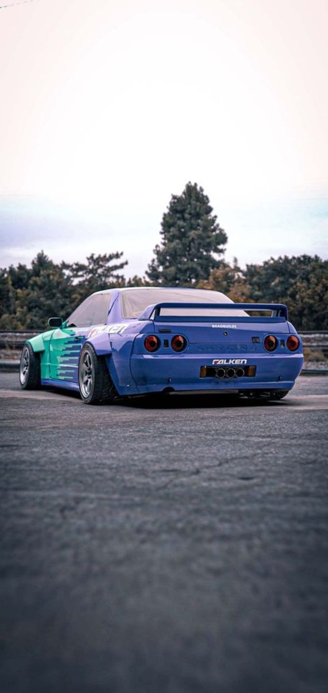 Nissan Skyline R32 with green and blue widebody kit and rear spoiler parked in front of overcast trees.