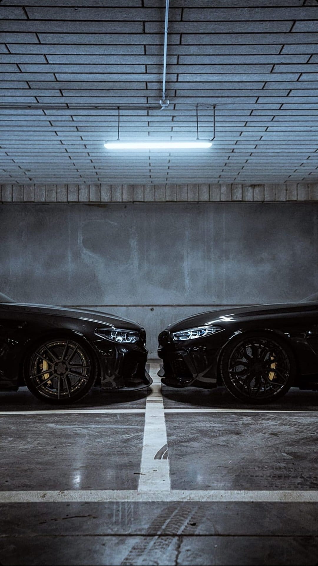 Two black luxury sports cars parked head-to-head in a dark garage with a blue fluorescent overhead light glow.