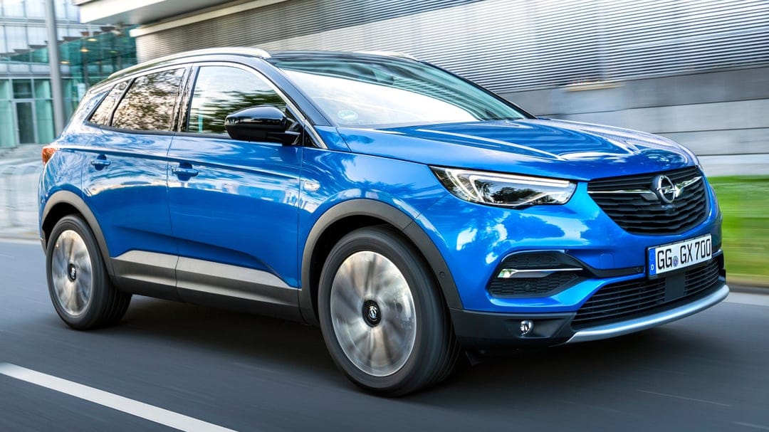 Blue Opel Grandland X SUV in motion on a road with a metallic building background and motion-blurred wheels.