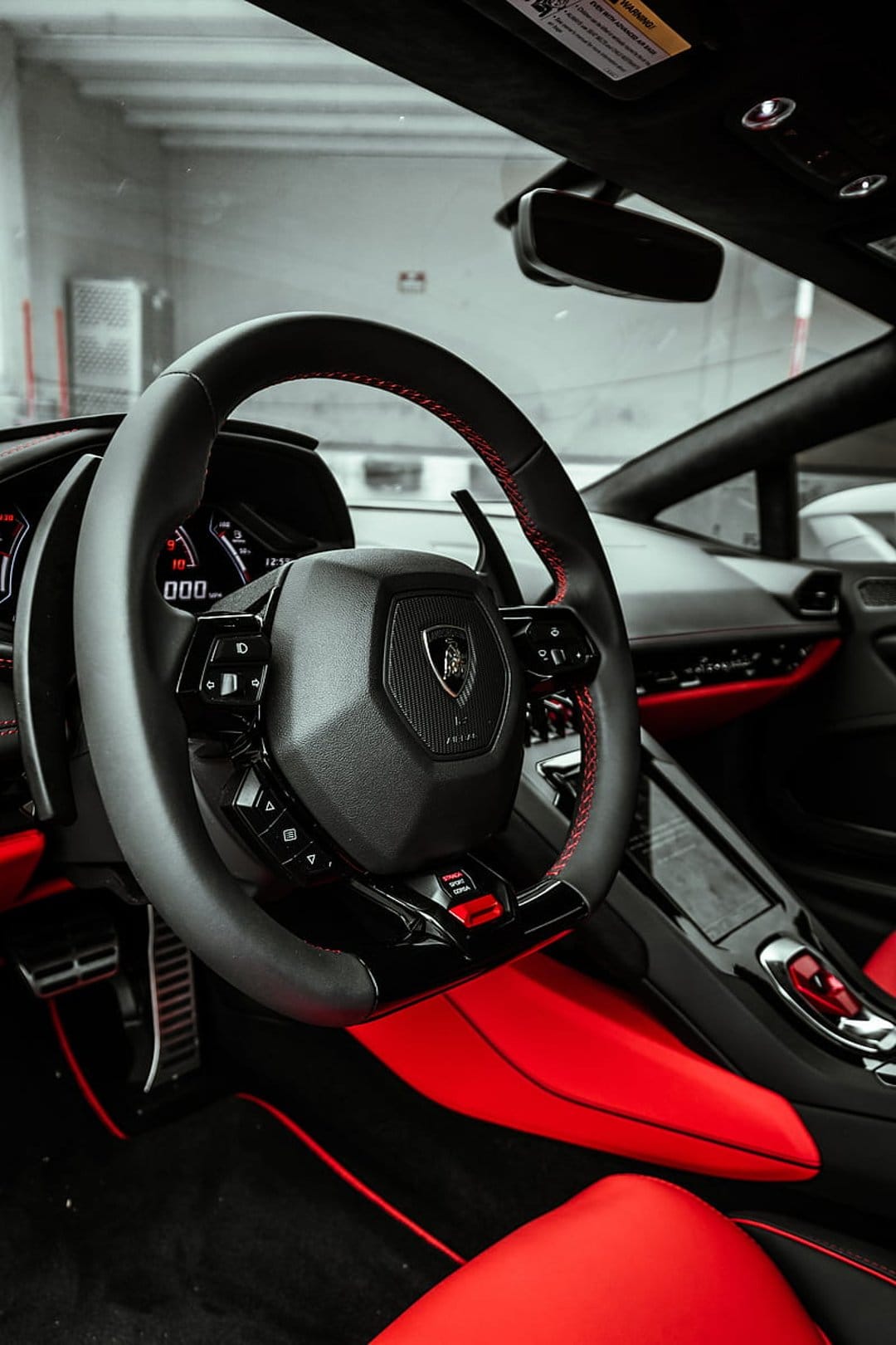 Lamborghini steering wheel with red stitching and emblem inside a black and crimson luxury supercar cockpit.