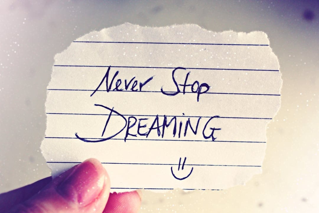 Fingers hold a torn paper note with handwritten Never Stop Dreaming text over a blurred sparkling background.