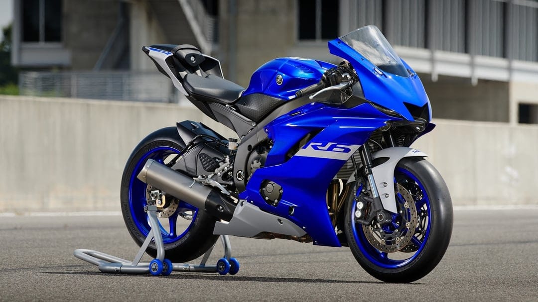 Blue Yamaha R6 motorcycle on a rear paddock stand at a racetrack with concrete walls and building background.