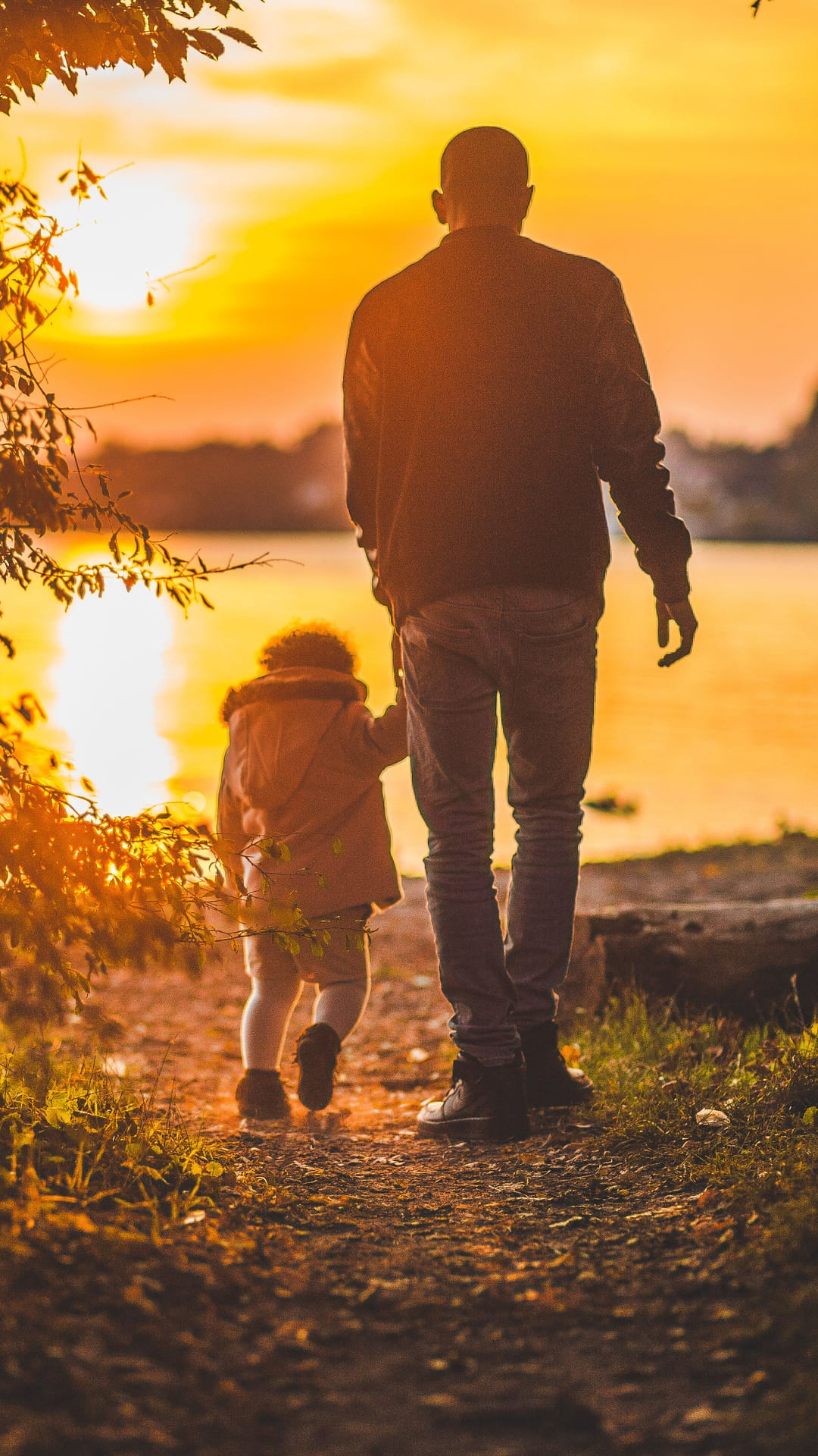 Parent and child silhouettes walk hand-in-hand along a glowing lakeside path during a vibrant golden sunset.