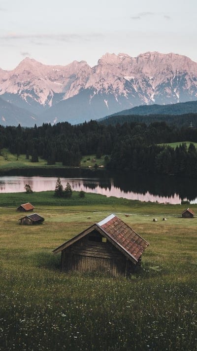 This serene vertical landscape captures a peaceful countryside scene with scattered wooden barns nestled in lush green meadows. A calm lake reflects the dramatic, snow-dusted mountains that rise majestically in the background. A dense forest line separates the meadow from the lake, and the soft light of dusk or dawn casts gentle hues across the sky, creating a tranquil and picturesque setting.