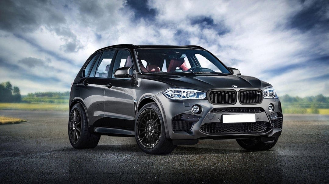 Gray BMW X5 M performance SUV with red seats and black wheels parked on a wet road under cloudy skies.