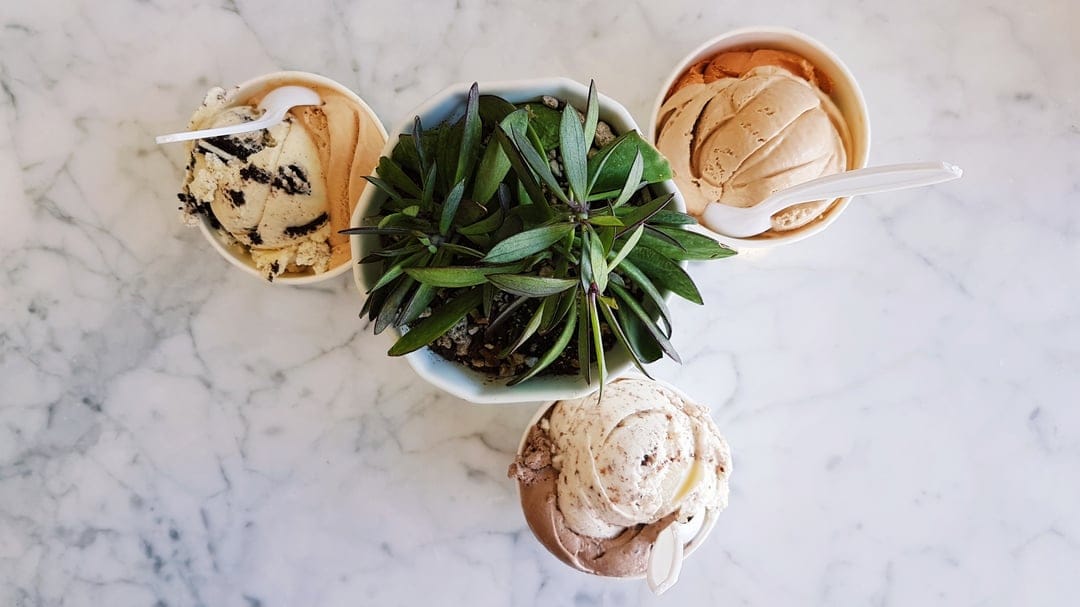 Three ice cream cups and a small green plant on white marble in a top-down food photography composition.