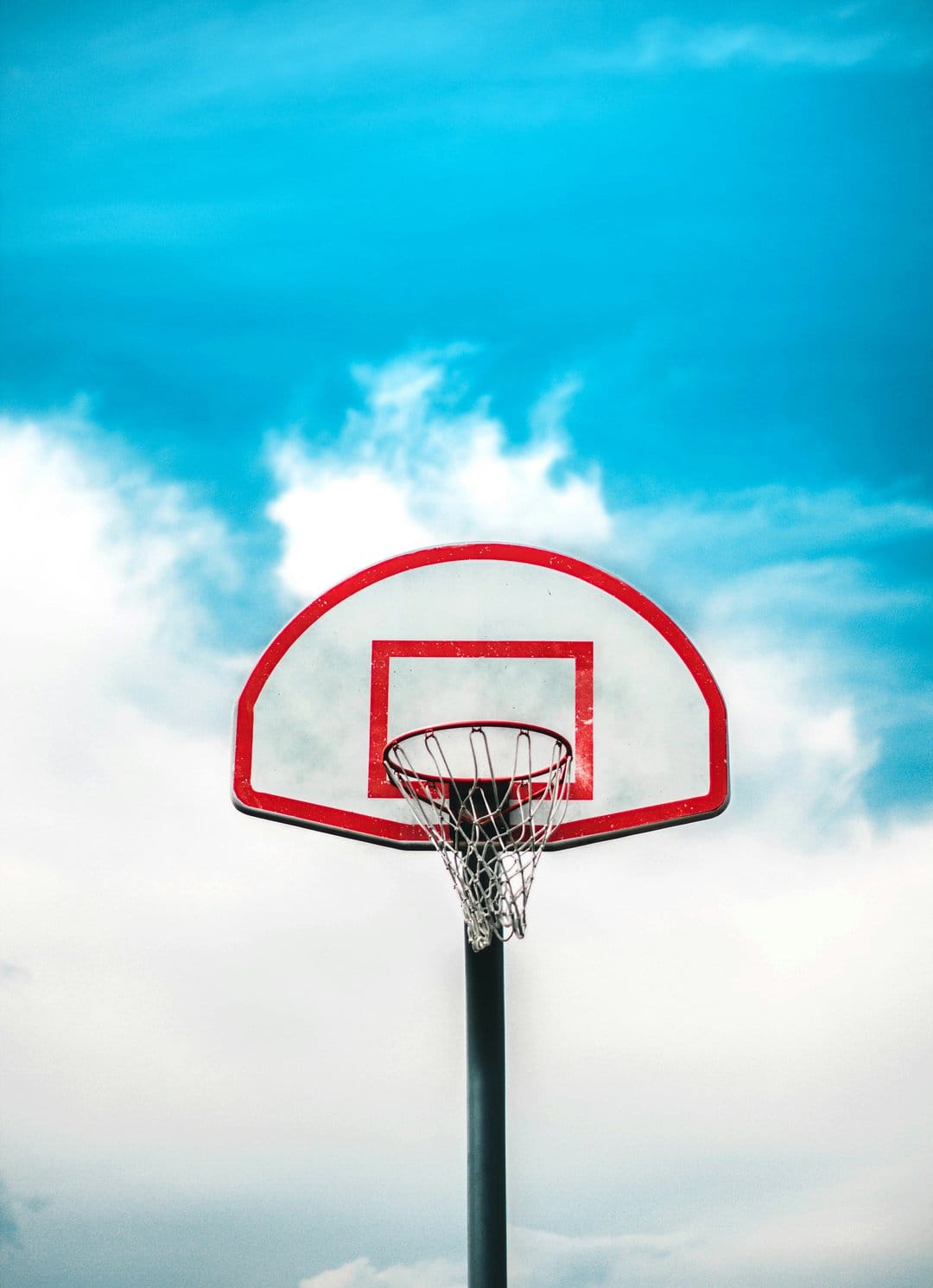 Basketball hoop with a red rim and white backboard set against a bright blue sky with wispy white clouds.