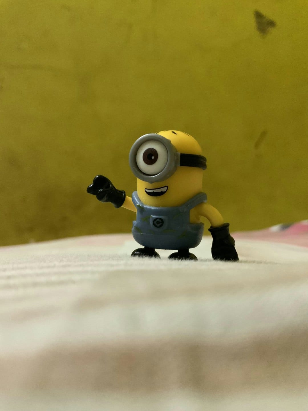 One-eyed Minion toy wearing blue overalls waves a hand while standing on a white and yellow textured background.