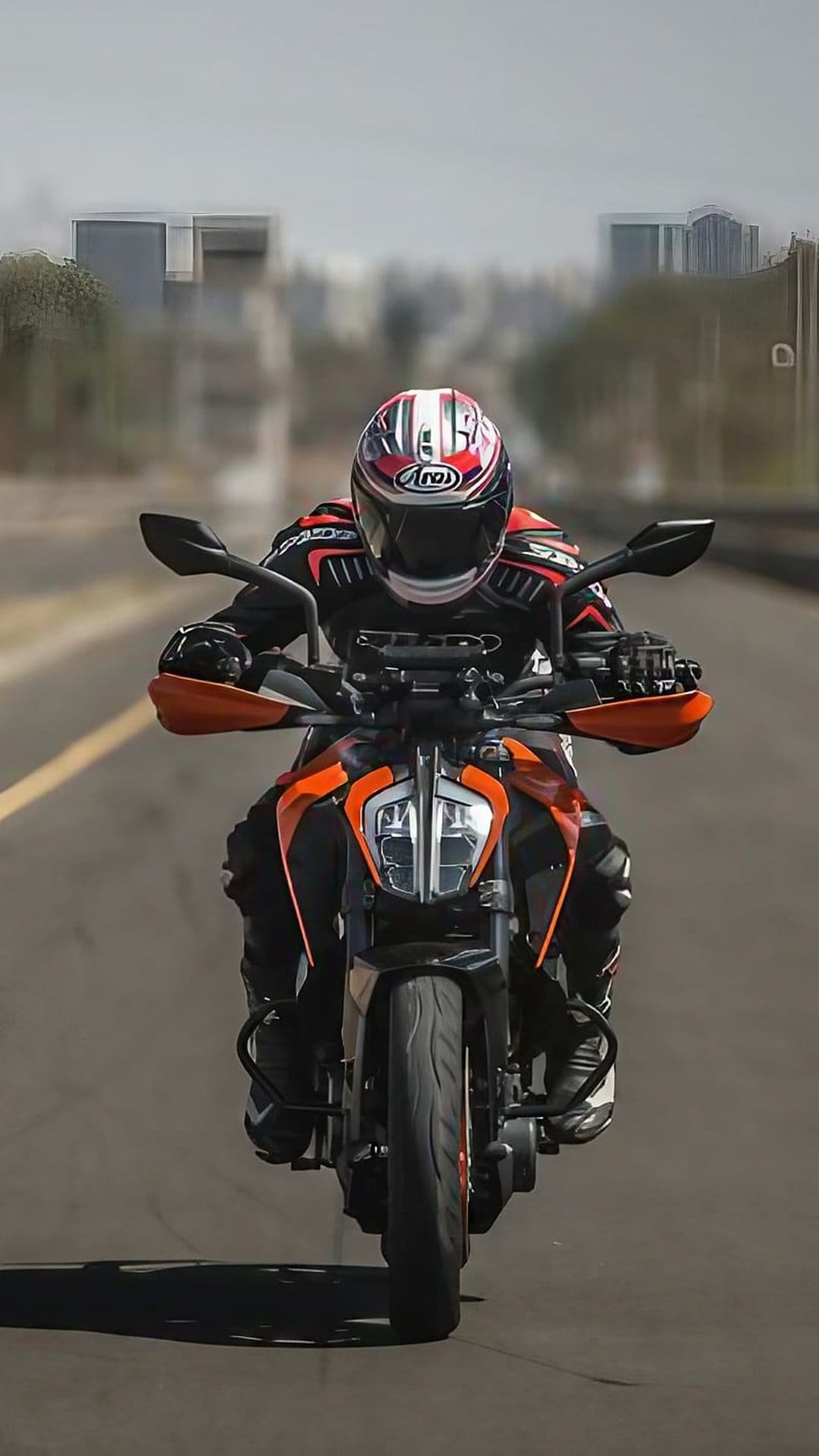 Motorcyclist in a red and white Arai helmet rides an orange KTM bike past blurred city buildings on a paved road.
