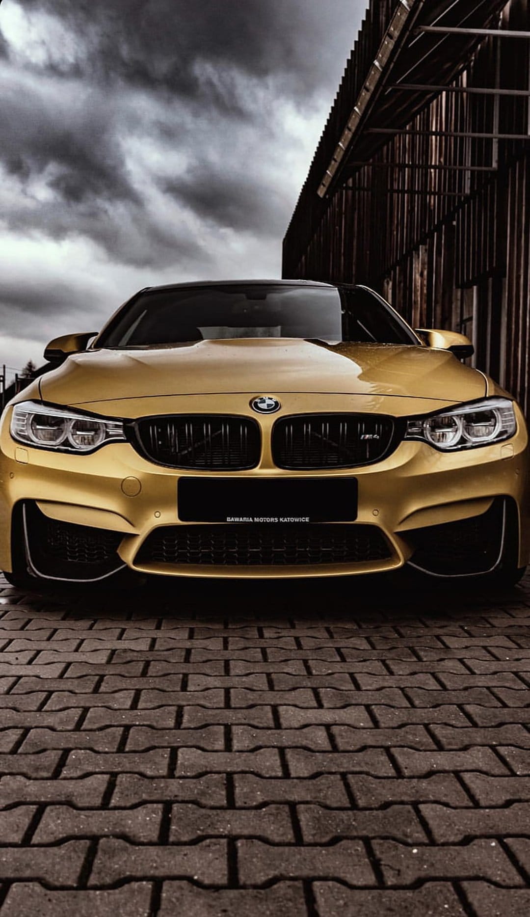 Golden BMW M4 parked on a brick road under a dramatic cloudy sky with its iconic headlights and grille visible.