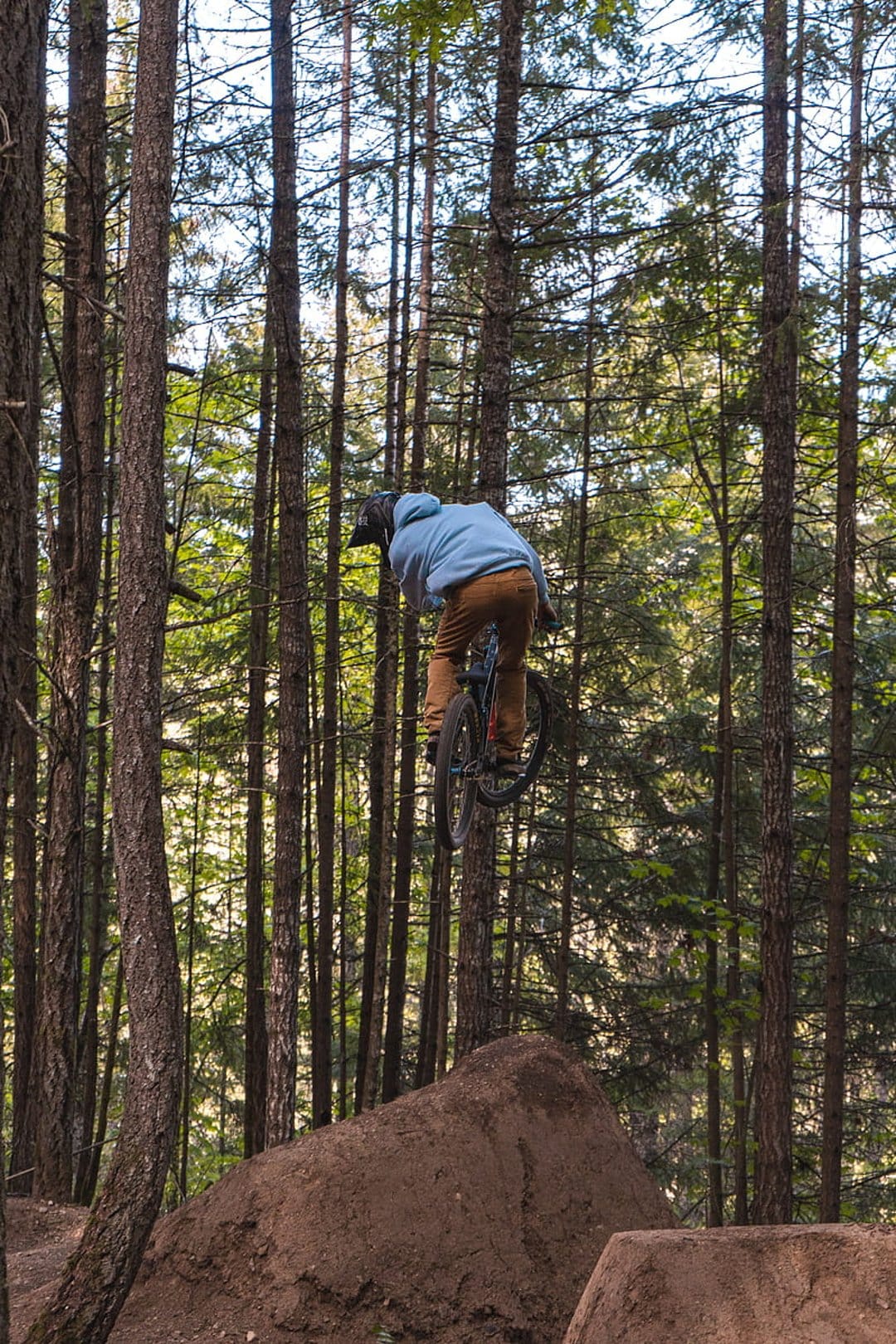 Mountain biker in blue gear performs a high-air dirt jump surrounded by towering trees in a dense forest.