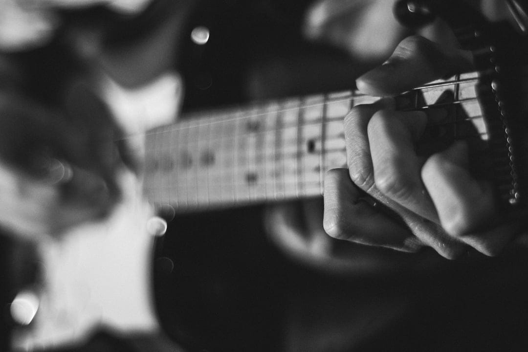 Musician's fingers press strings on an electric guitar fretboard in a high-contrast monochrome close-up.
