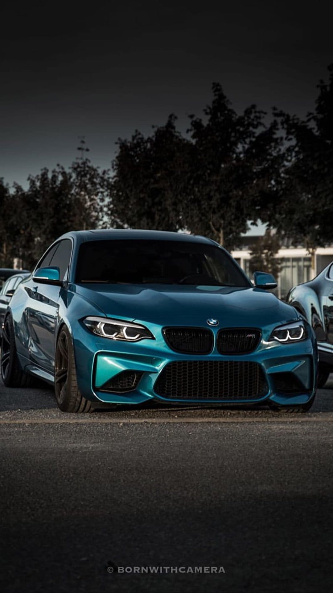 Teal BMW M2 coupe parked on asphalt with aggressive front styling and a dark treeline background under a moody sky.