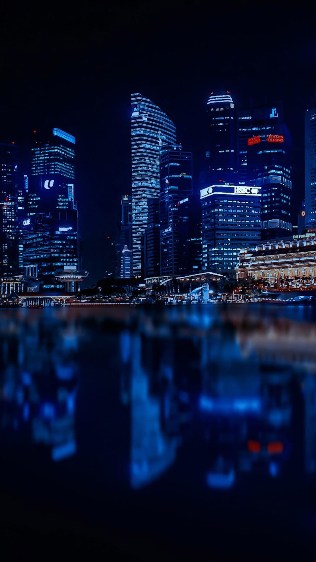 Blue illuminated skyscrapers reflected in calm harbor water under a dark night sky for mobile backgrounds.