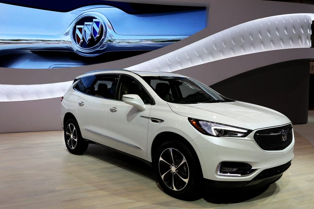 White Buick Enclave SUV on a light wood floor with a large Buick logo background in a modern showroom.