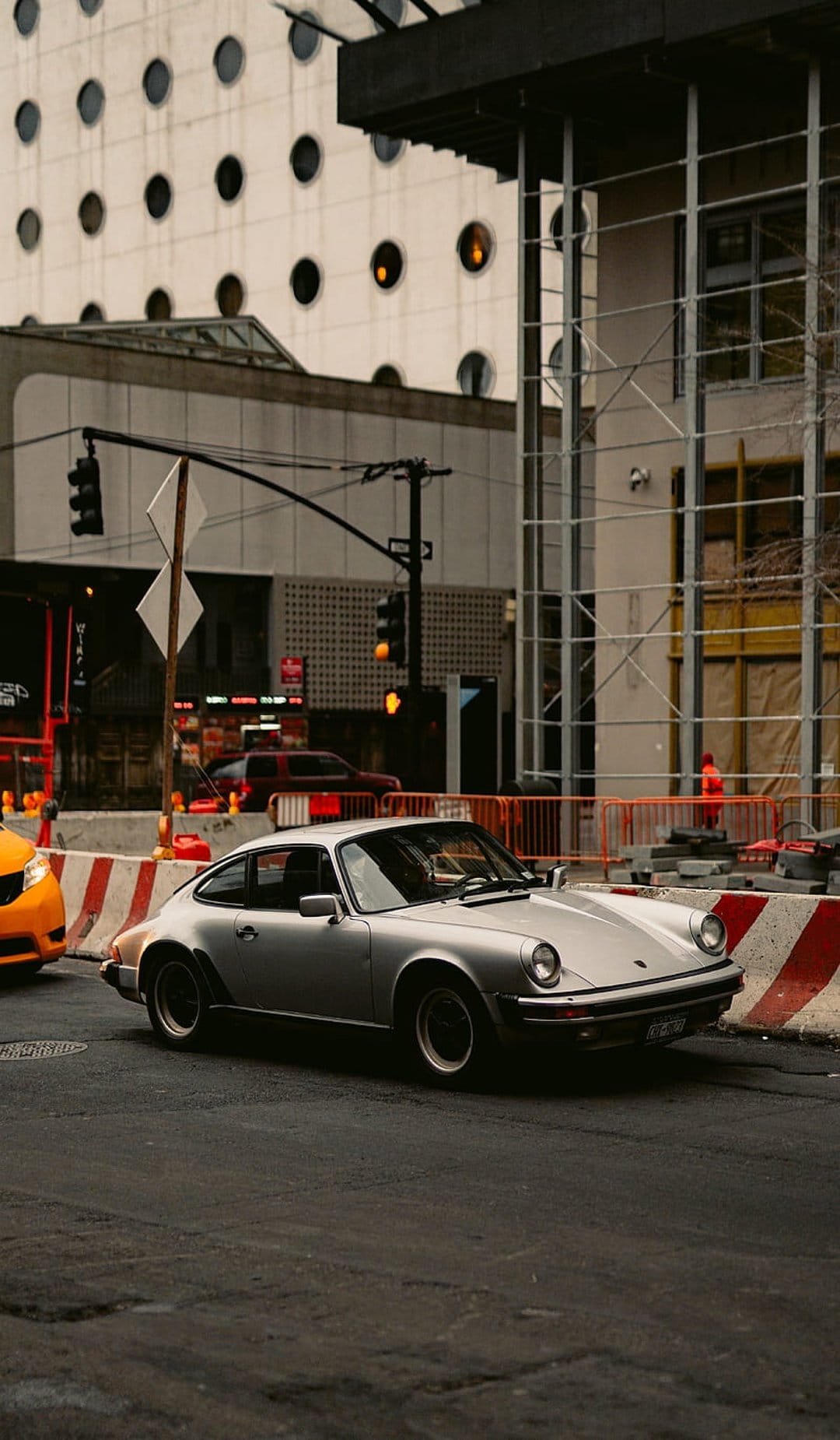 Silver Porsche 911 drives on a city street past orange construction barriers and scaffolding under a tall building.