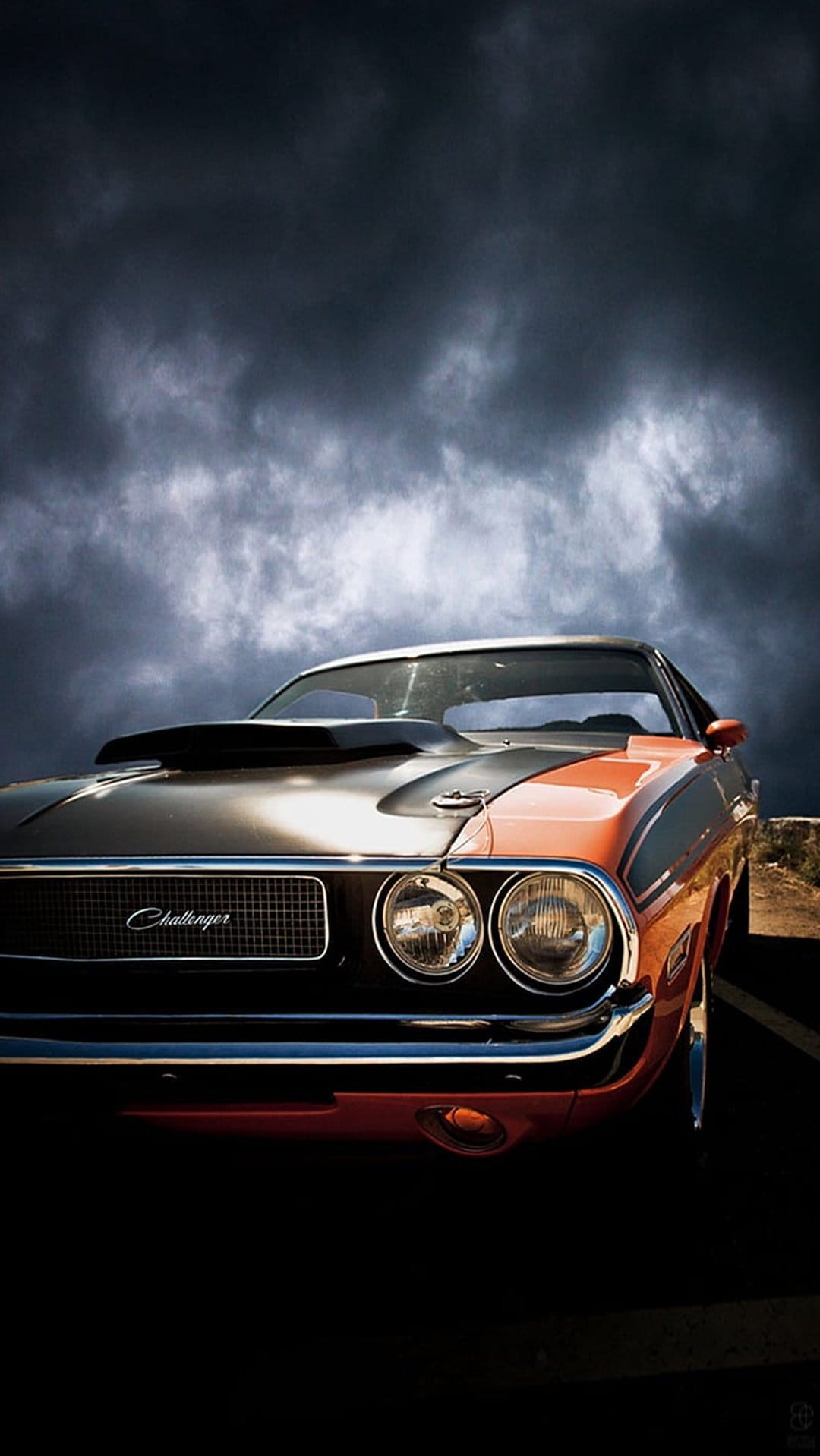 Orange Dodge Challenger muscle car with black hood scoop parked under a dark stormy sky with glowing headlights.