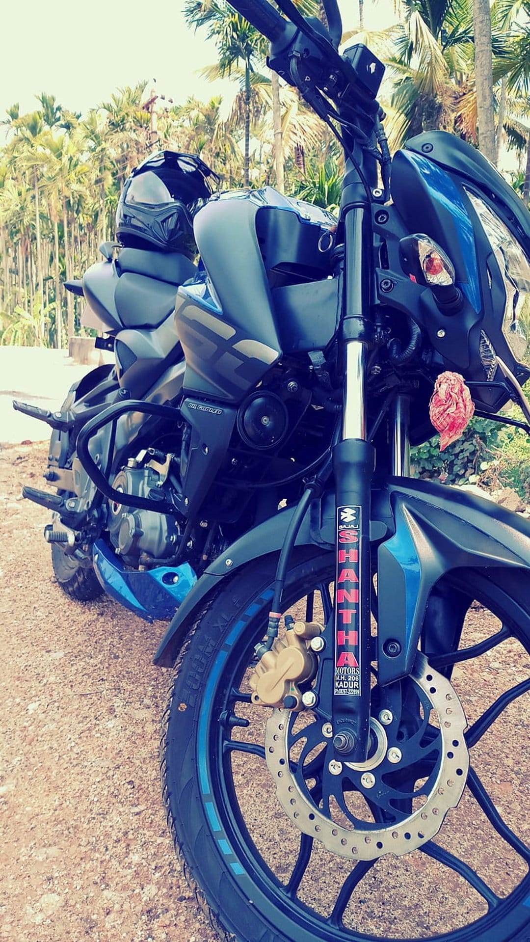 Blue sport motorcycle with a black helmet on the seat parked on a dirt path with blurred palm trees behind.