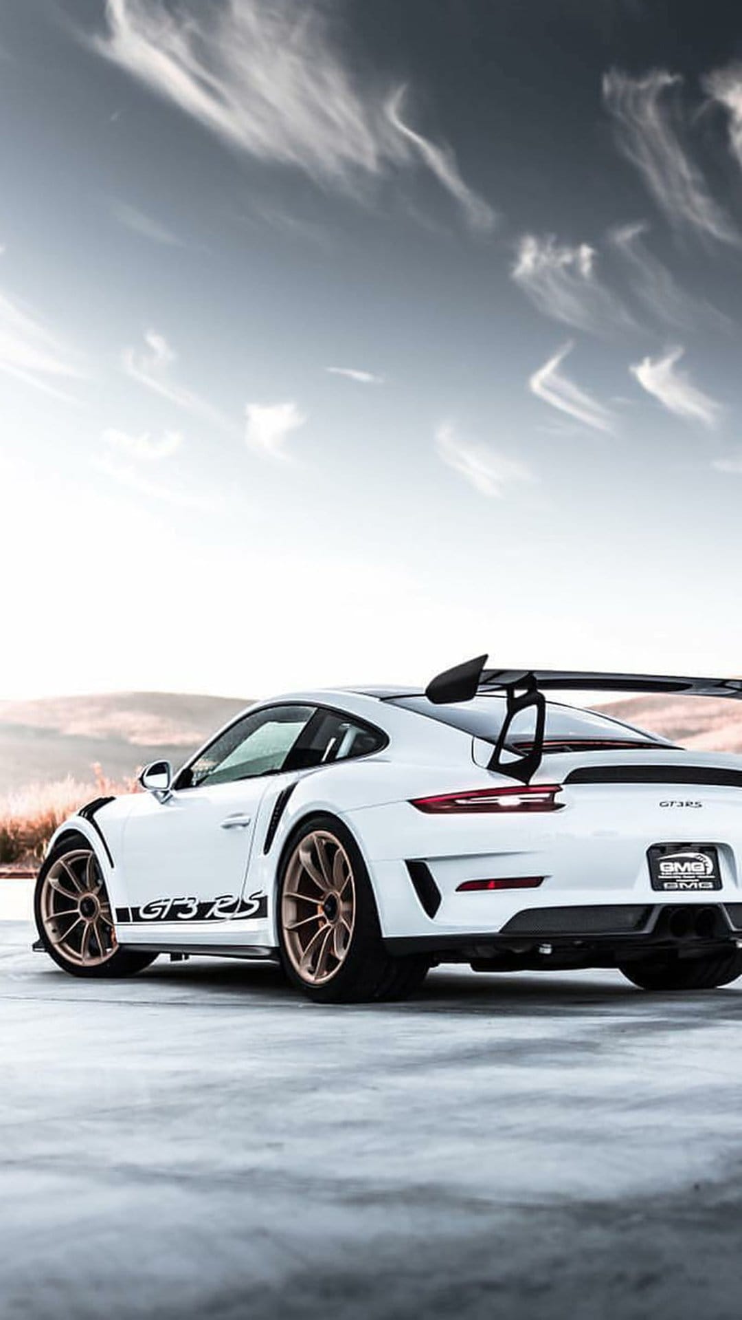 White Porsche GT3 RS showing a rear wing and gold wheels on a road under a wispy sky with hills in distance.