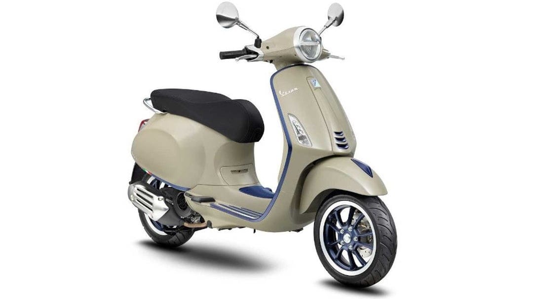 Beige Vespa Primavera scooter with blue rims and chrome mirrors on a white studio background with soft shadows.