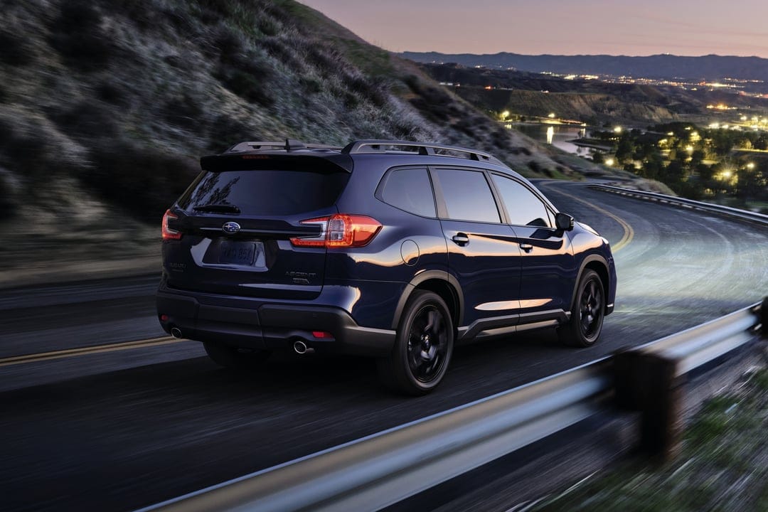Blue Subaru Ascent driving on a winding road at dusk with city lights reflecting in the valley background.