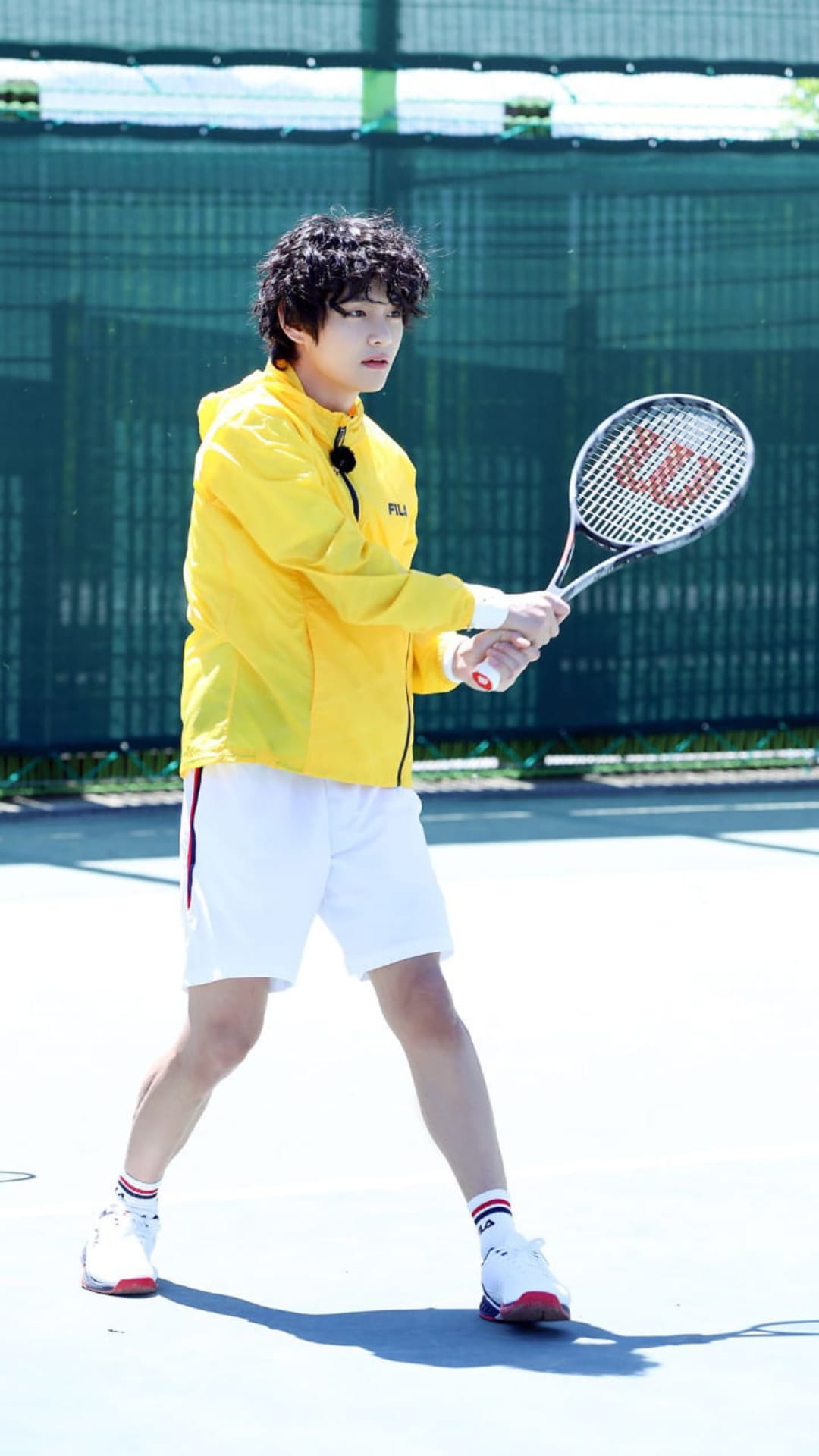 Kim Taehyung holds a tennis racket on a court wearing a bright yellow jacket and white athletic shorts in sunlight.