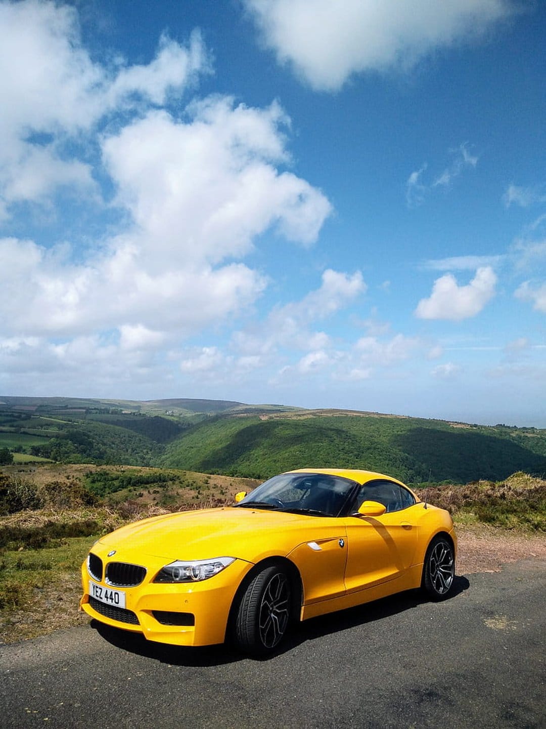 Yellow BMW Z4 convertible parked on a winding asphalt road overlooking a lush green valley under a blue sky.