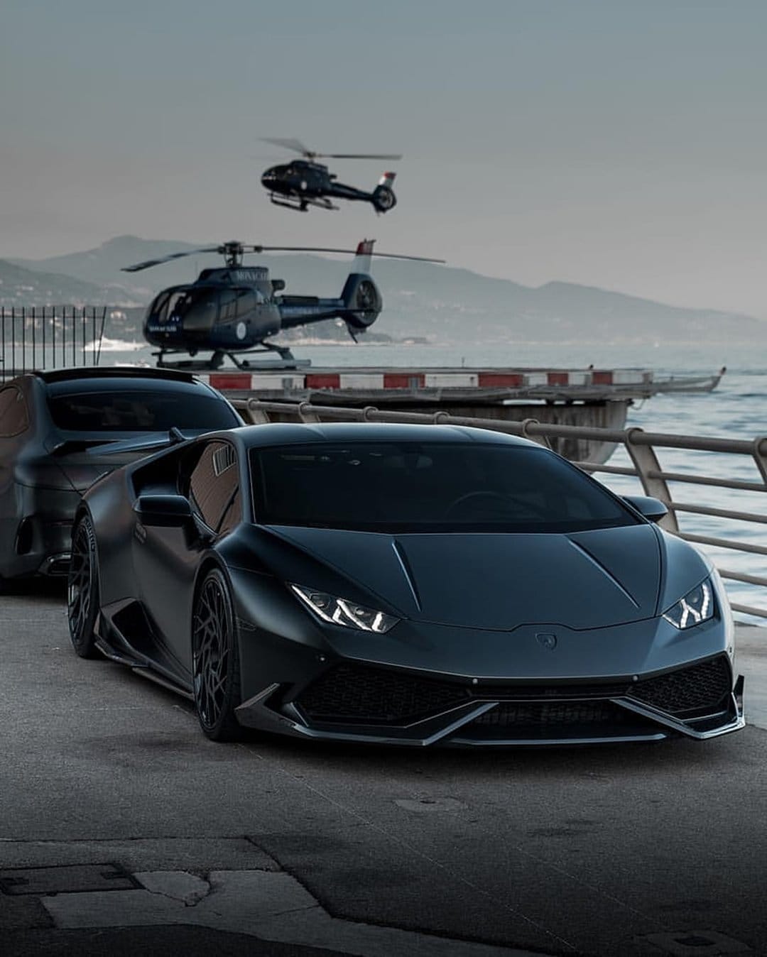 Matte black Lamborghini and luxury SUV parked by a misty sea with two helicopters flying over coastal mountains.