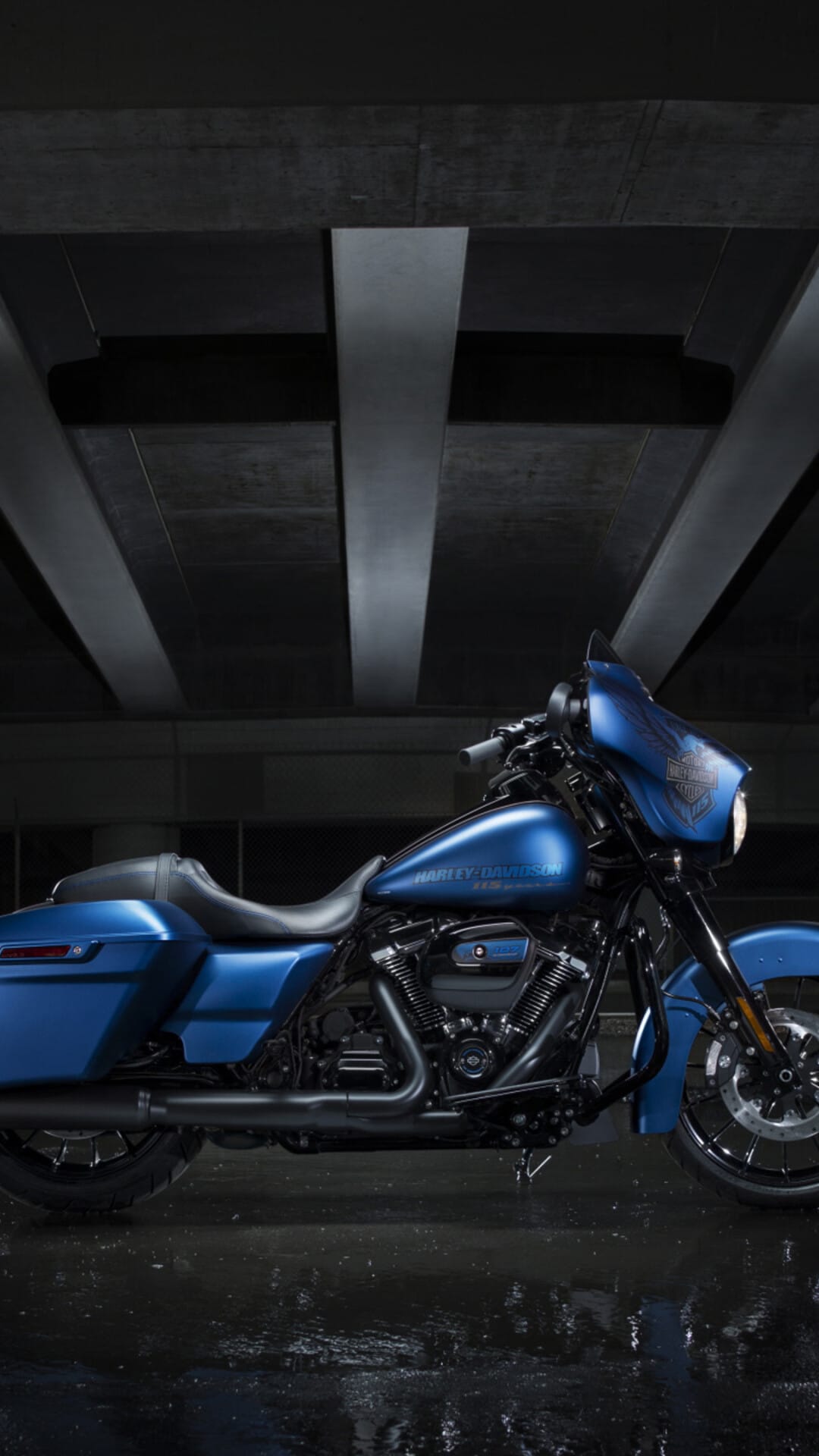 Blue Harley-Davidson motorcycle parked under concrete pillars with reflections on a wet ground.