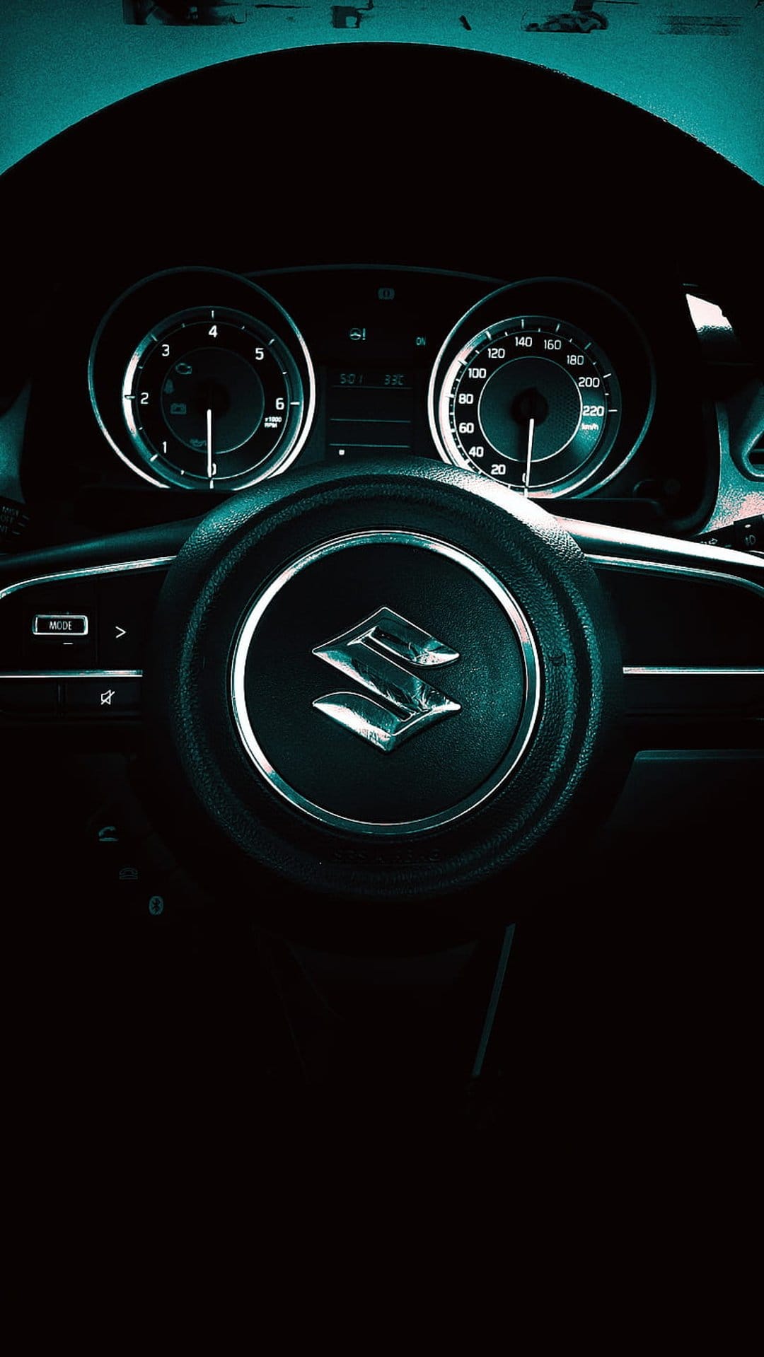 Suzuki steering wheel emblem and illuminated teal dashboard gauges captured in a close-up interior night shot.