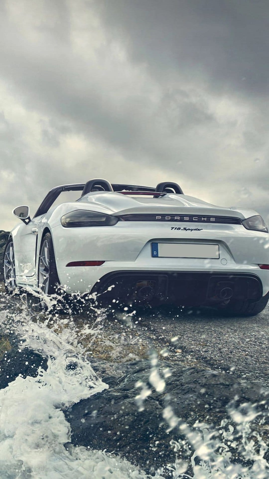 White Porsche 718 Spyder rear view splashing through blue ocean waves under a dramatic and cloudy grey sky.