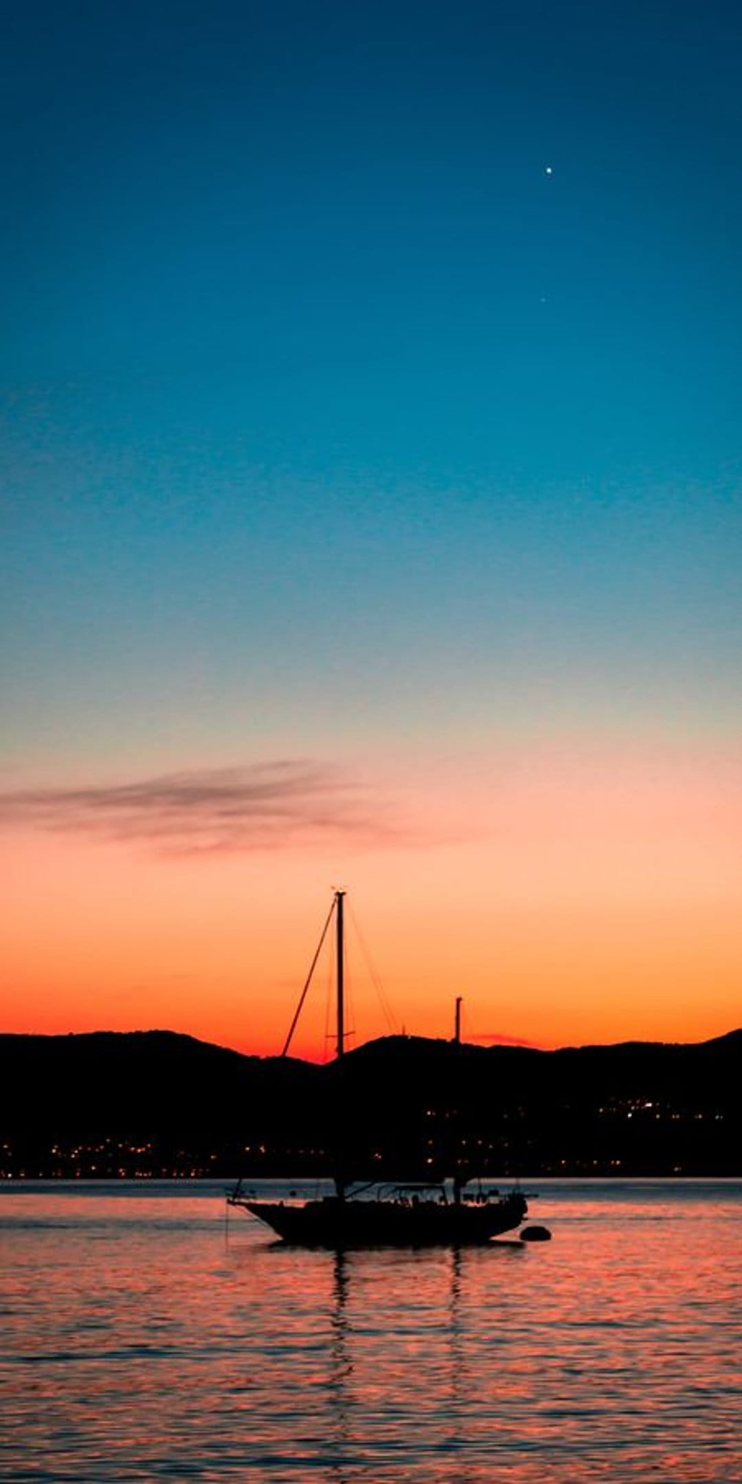 Sailboat silhouette on calm water under a red sunset sky with a single star and distant glowing city lights.
